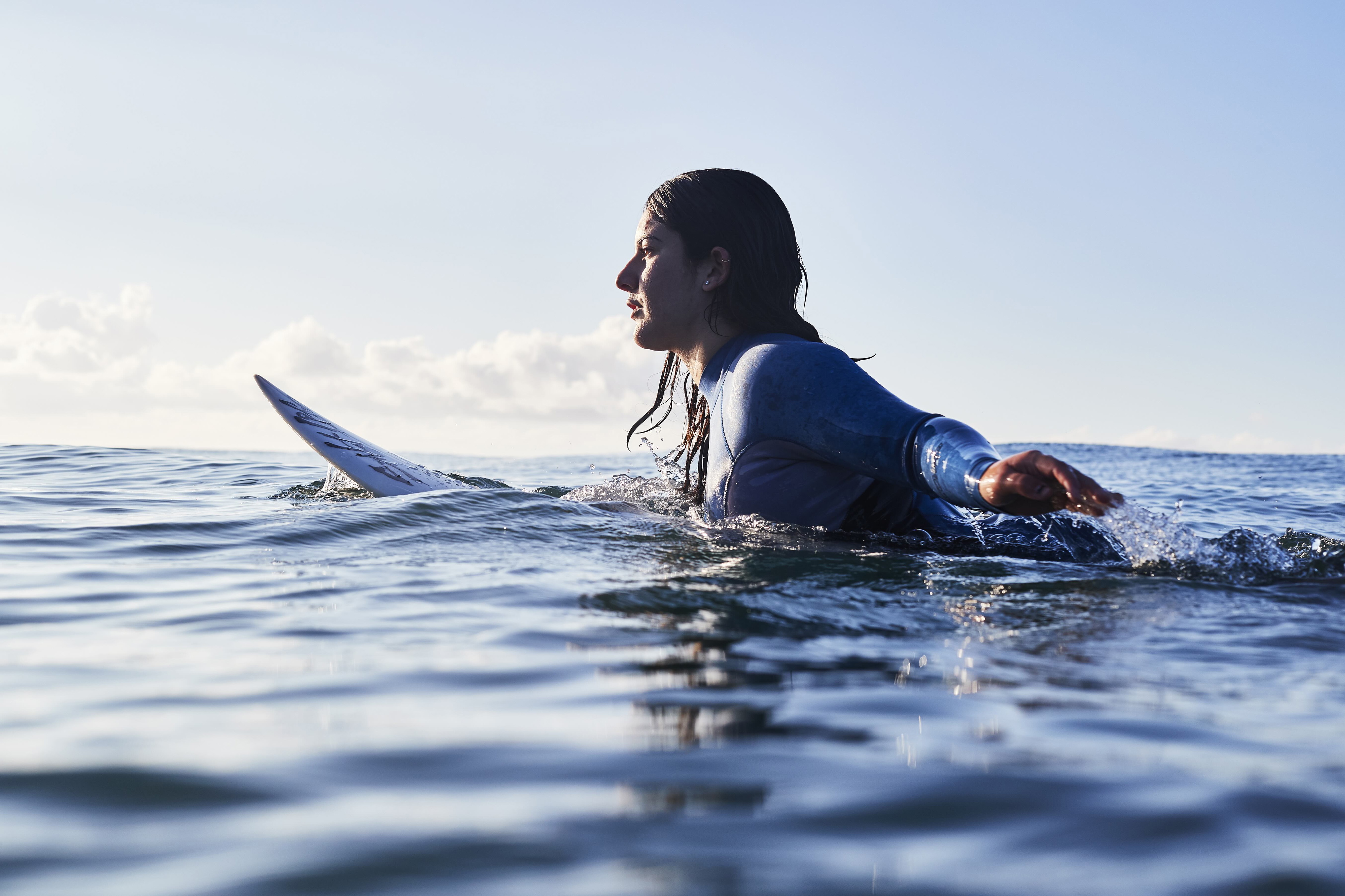 Woman in a blue-sleeved wetsuit resting on her surfboard in calm open water, looking along the horizon