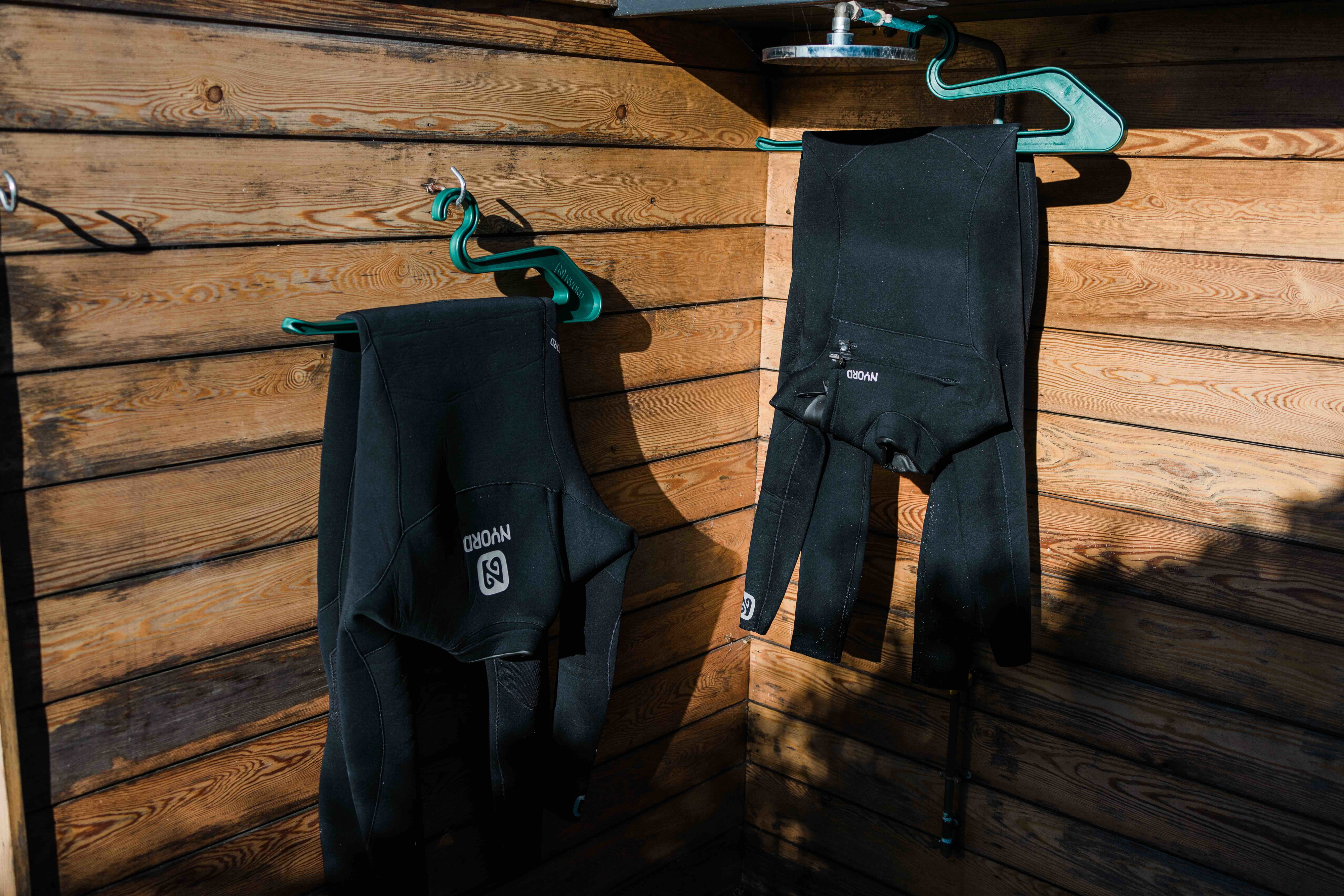Two black wetsuits hung on hangers to dry under a wooden beach shelter