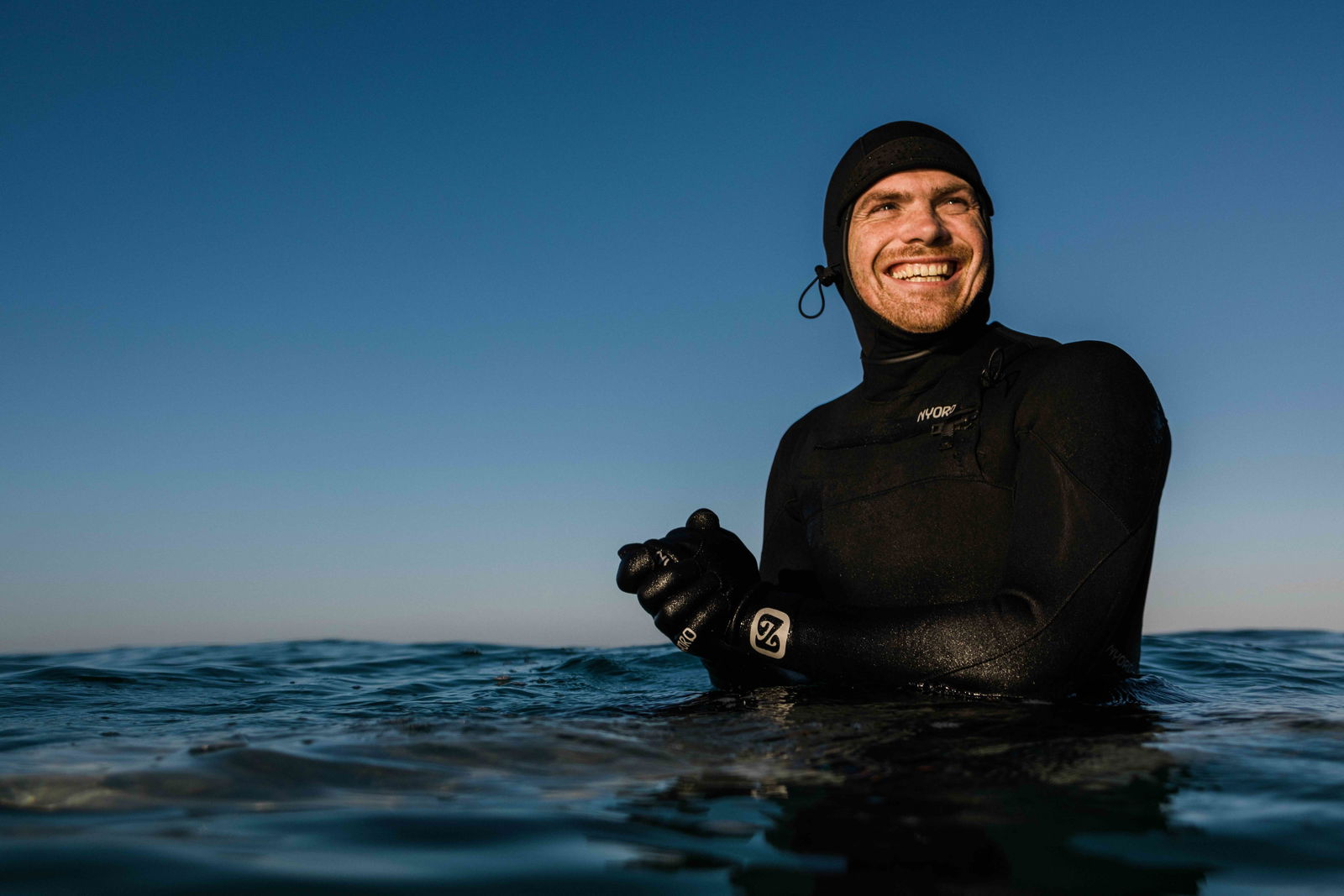 Smiling man in a hooded black wetsuit and gloves standing chest deep in the sea