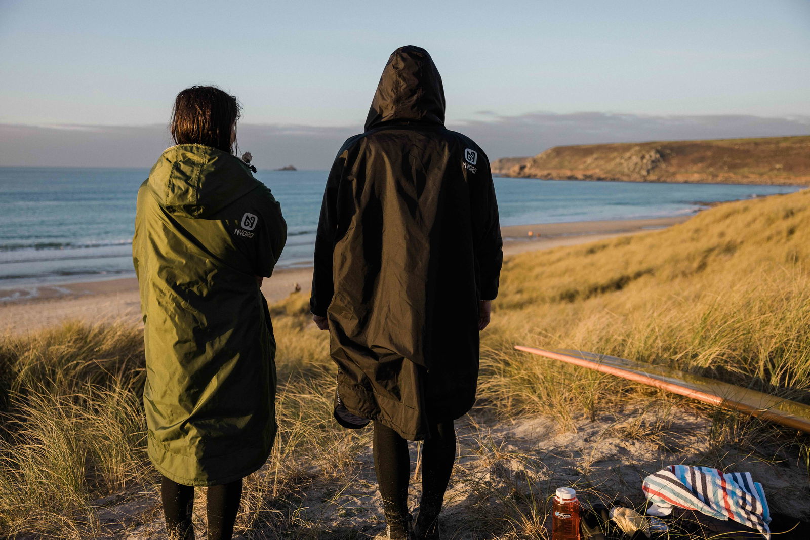 Two people in long hooded changing robes looking out over a beach at sunset