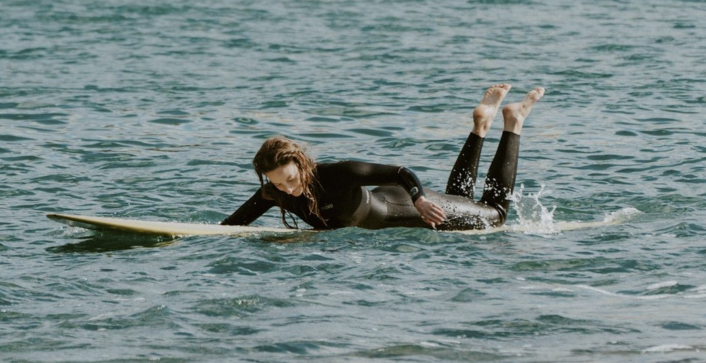 Two surfers in black wetsuits paddling out together on a single longboard