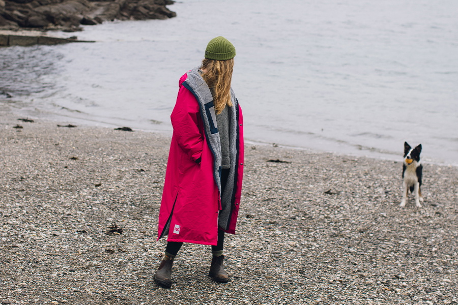 Woman in a pink and grey changing robe and green beanie standing on a pebble beach with her dog